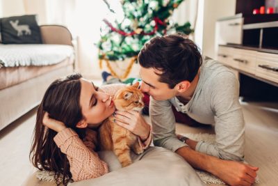 A man and woman kissing their cat on the floor of a living room in front of their Christmas tree