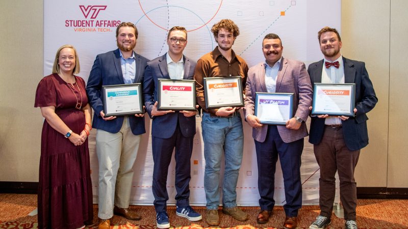 Six people stand smiling, the five on the right holding award certificates.